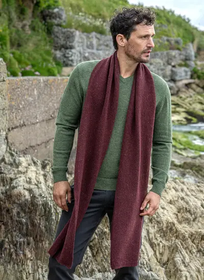 Man wearing a burgundy wool cashmere tweed scarf draped over a green sweater, standing on a rocky coastal path in Ireland.
